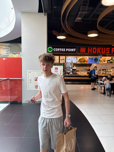 The image shows a young man standing in what appears to be a food court or shopping mall. He is wearing a white t-shirt and grey shorts and is holding a smartphone in his right hand and a brown paper bag in his left. Behind him, a food establishment with signage that reads "COFFEE POINT HOKUS POKUS" is visible. The establishment has menu boards displaying food items and prices, including an offer for "HAPPY HOURS -50%". Other people are seen in the background, some seated at tables. The overall setting suggests a casual, indoor environment. The lighting appears to be artificial, consistent with an indoor location. The floor is tiled in a black and white pattern. The image is oriented vertically.