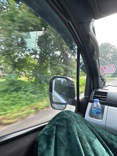 The image shows the interior of a vehicle in motion, viewed from the passenger's perspective. Lush green foliage and trees blur outside the windows, indicating a rural or suburban environment. Sunlight filters through the trees, creating a bright, daytime scene.
Inside the car, a dark green blanket is draped over the passenger's legs. A side mirror is visible, reflecting the outdoor scenery. The dashboard features an air vent and a cup holder containing a bottle of Nestlé water. A red sticker with text in Thai is affixed to the windshield. The vehicle appears to be traveling on a paved road, with evidence of passing buildings and utility poles visible in the distance.
The specific location is identified as Ban Bang Hin, Thailand. This suggests the vehicle is traversing a road within or near this community. The overall impression is one of a peaceful journey through a verdant landscape in Thailand.