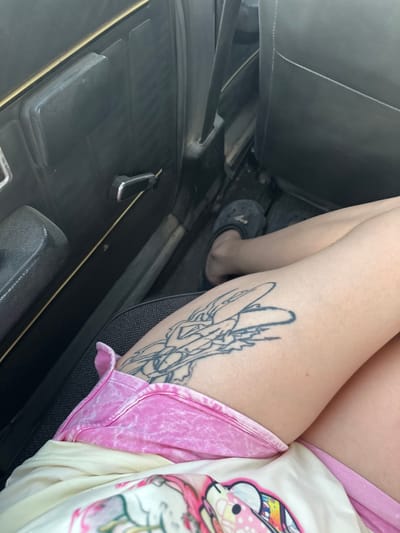 The image shows a close-up view of a person's leg with a tattoo, sitting inside a vehicle. The person is wearing pink shorts and a white shirt with a colorful cartoon-like graphic on it. The tattoo on the leg appears to be a stylized drawing of a mythical creature with wings.
The interior of the car is visible, including a dark-colored seat, part of the dashboard, and a handle on the door. The overall lighting suggests it is daytime, but the specific time or weather conditions are not discernible. The presence of the car's interior indicates the scene is set within a vehicle, likely in motion or parked. The focus on the person's leg and clothing, along with the tattoo, suggests a casual and personal moment. No visible text is present in the image.