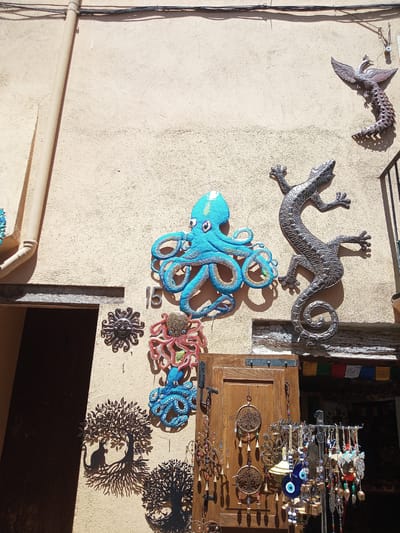 The image depicts a colorful and eclectic display of decorative items mounted on a textured, beige stucco wall in the historic town of Besalú, Spain. Dominating the upper portion of the wall are several metallic and ceramic sculptures. A large, vibrant blue ceramic octopus with pink eyes is prominently featured, positioned to the left of a life-sized, intricate metallic sculpture of a lizard with a coiled tail. To the upper right, a metallic sculpture of a phoenix-like bird perches on a coiled, serpentine body. Below these larger pieces, smaller decorative elements are visible. These include a sun-shaped metal ornament with a human-like face, a red ceramic octopus, and two silhouette-style metal sculptures of trees, one featuring a cat perched on a branch.

The wall itself shows signs of age, with visible cracks and imperfections. A beige pipe runs vertically along the left side of the wall, casting a shadow. To the left of the pipe, a dark doorway leads into a building. Below the sculptures, a weathered wooden door is visible, adorned with several hanging ornaments. These include two circular tree-of-life metal decorations, several strings of beads, bells, and protective amulets, such as blue and white "evil eye" talismans. A rack of similar charms and trinkets is displayed in front of the door, suggesting a market stall or a shop. Colorful prayer flags are strung across the top of the doorway. The overall impression is one of an outdoor marketplace or a quaint shopfront, with an emphasis on artisanal crafts and cultural motifs. The bright sunlight suggests it is daytime, with strong shadows cast by the objects and architectural features.