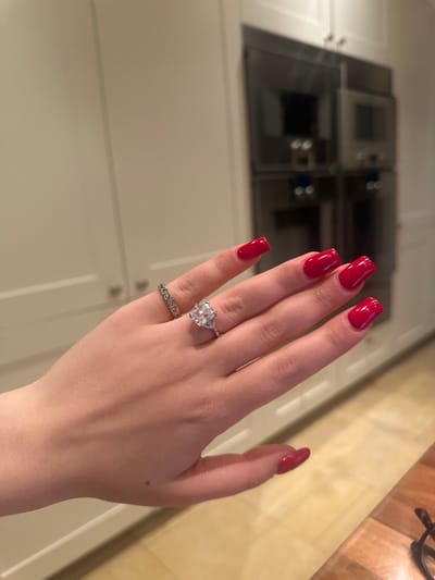 A hand displays red square-shaped nails and two rings, one with a large, ornate stone, the other a silver band with small, square settings. The hand is positioned against a background of white kitchen cabinets and stainless steel ovens. A countertop runs along the right edge of the frame, featuring a warm, wood-like surface.