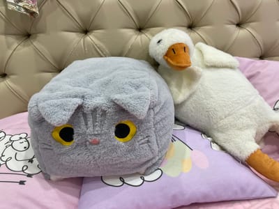 A gray, cat-shaped plush pillow and a white, duck-shaped plush toy rest on a pink and purple pillow adorned with drawings of sheep and clouds. The backdrop is a padded, beige headboard. The location of the scene is not discernible from the image.