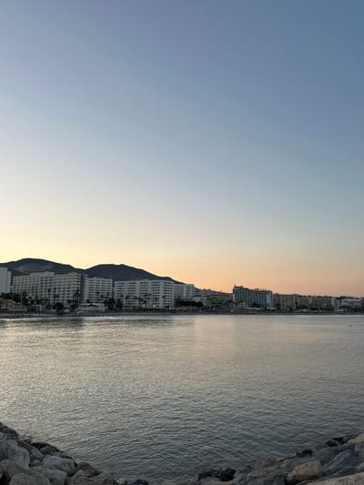 The image shows a coastal scene in Arroyo de la Miel-Benalmadena Costa, Spain, at either sunrise or sunset. The sky is a gradient of pale blue to soft orange. In the middle ground, a long stretch of white buildings, likely hotels or apartment complexes, lines the coastline, backed by dark, low-lying hills. Palm trees are visible in front of some of the buildings. The foreground is dominated by the sea, which is calm and reflects the colors of the sky. Rocky breakwaters are visible in the lower corners of the image. A few indistinct figures can be seen on a beach in the distance, along with some beach umbrellas and loungers. The overall impression is a tranquil, developed coastal resort area.