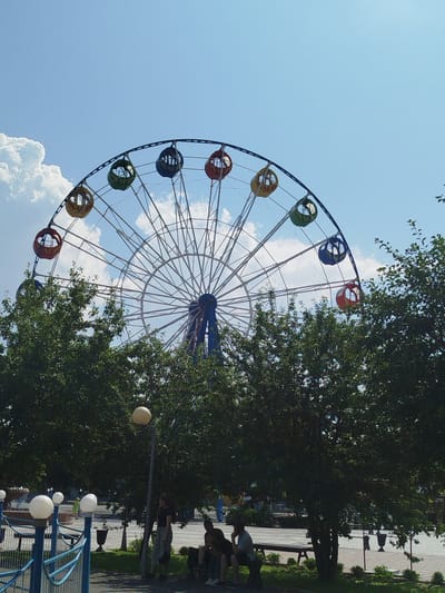 The image shows a Ferris wheel in an amusement park. The Ferris wheel has a white structure with colorful gondolas. In the foreground, there are trees and people sitting on benches. The sky is blue with some clouds. This Ferris wheel is located in the city of Yalutorovsk, Russia.