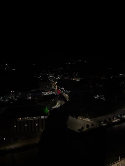 The image depicts a nighttime aerial view of the city of Kapan, Armenia. The scene is characterized by darkness punctuated by illuminated buildings and streetlights, creating a stark contrast between light and shadow. In the foreground, a large, dark shadow, likely from a person or structure, obscures a significant portion of the view, casting an almost silhouette-like effect on the buildings below.

Several multi-story buildings are visible, with windows lit from within, suggesting occupancy. One prominent building on the left side of the frame features vertical stripes and spotlights illuminating its facade. Further into the cityscape, another taller building displays a red "HOTEL" sign. Roads and vehicles are sparsely discernible amidst the darkness, with headlights and street lamps casting pools of light. The surrounding landscape appears to be hilly or mountainous, with distant lights indicating the presence of more structures further out. The overall impression is one of a quiet, nocturnal urban environment.