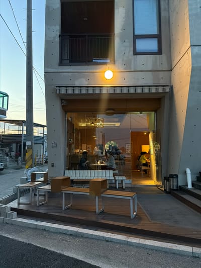 The image shows a commercial building in Toseong-myeon, South Korea, featuring a cafe with a glass storefront. Patrons are visible inside. Outdoor seating, including wooden benches, is placed on a raised wooden platform next to the sidewalk. The building is made of gray concrete, with two windows on the upper floors. Electrical lines and poles are seen in the foreground. The sky is visible, suggesting daytime.