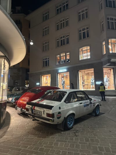 A nighttime street scene in Sankt Moritz, Switzerland, displays four vintage rally cars parked sequentially on a cobblestone street. In the foreground right, a white classic rally car, resembling a Ford Escort RS2000, features the number '57', 'Lombard Rally', 'VREDESTEIN TYRES', and 'Castrol' decals, blue multi-spoke wheels, a visible roll cage, and a Swiss license plate 'AH OA 9H'. Behind it is a red classic car, likely a Fiat 500, with a '32' decal and partial license plate 'BOR OA 7', followed by a dark blue classic car, resembling a VW Beetle, numbered '12', with a partial license plate 'PJK 153 929'. The last visible vehicle is a dark green classic sedan.

The cars are positioned in front of a multi-story building with a light-colored facade. The ground floor of this building houses an illuminated PRADA luxury fashion store, featuring large display windows showcasing mannequins dressed in apparel. Above the storefront, the building has multiple arched and rectangular windows. One individual wearing a yellow reflective vest stands on the cobblestone pavement near the PRADA store, facing away from the camera. Several other people are visible inside the PRADA store. To the left, a building with a curved, light-colored architectural overhang partially obscures a sign that reads "wega Buchhandlung Papeterie Souvenirs". Overhead street lighting provides additional illumination.
