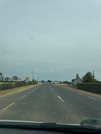 The image shows a view from a vehicle traveling on a rural road in Killcommon, Ireland. The road is paved with asphalt and has yellow dashed lines indicating traffic lanes. There are white dashed lines on the right side of the road. The sky is overcast with light gray clouds.
On the left side of the road, there is a field of dry grass and a row of hedges. Several houses with pitched roofs are visible in the distance. A yellow traffic sign with a black symbol and "100 m" written on it is placed on a pole on the side of the road. Further down the road, there are more houses and trees, with a stone wall and hedge lining the right side of the road. A few cars are visible on the road ahead. The foreground shows the dashboard and windshield of the vehicle from which the photo was taken.