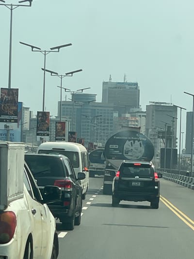 The image displays an elevated roadway in Ebute-Metta, Lagos, Nigeria, under a hazy, light grey sky. Traffic is visible across at least two lanes, with vehicles appearing stationary or slow-moving.
In the left foreground, the partial front-left section of a white vehicle is visible. Directly behind it is the rear of a dark-colored SUV, followed by the rear of a white minibus, and then another dark-colored SUV. In the adjacent lane to the right, a large tanker truck is prominent, with "PROTECTION in EVERY DROP" and "API SP" text, along with two lubricant bottles, displayed on its cylindrical tank. Another tanker truck is visible behind it. The roadway features double yellow lines on the right and dashed white lines marking lane divisions. Concrete barriers line the right edge of the roadway.
Numerous tall street lamp posts, each fitted with multiple horizontal luminaires, are positioned along the road. These posts also support large vertical advertising banners. One banner clearly promotes a film titled "COLOURS OF RAE," indicating "EXPERIENCE IN CINEMAS CHRISTMAS EVE 2023." Another banner features a distinct male portrait.
The background presents an urban skyline with multiple multi-story buildings. A prominent tall building with a grid-patterned facade is identifiable as the Union Bank of Nigeria Plc headquarters. To its right, another building with partially legible text, including "TRUST M," is visible. No individuals are observed outside of vehicles.