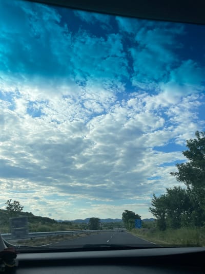 The image depicts a view from inside a vehicle traveling on a road, likely a highway, in Balabanovo, Bulgaria. The foreground shows the dashboard of the vehicle, obscuring the bottom portion of the frame. The road ahead is asphalt with white lane markings. To the right, there are trees and dense green vegetation. A blue road sign with white text and arrows is visible in the distance, indicating directions. The landscape beyond the road features rolling hills and some scattered buildings. The sky is partially cloudy with patches of blue. The overall scene suggests a daytime journey through a semi-rural or suburban area.