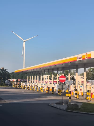 The image captures a Shell truck stop and gas station under a clear blue sky. In the background, a large white wind turbine with red tips stands tall against the sky. The gas station features a prominent yellow and white awning with the Shell logo and "TruckDiesel" branding. Rows of fuel pumps are visible under the awning, each with yellow and black bollards surrounding them. A red and white circular "no entry" sign is posted on a pole near the pumps. The ground is asphalt, with some grassy areas and rocks. The scene appears to be in a semi-rural or industrial area, possibly on the outskirts of a city. No people are visible in the image. The overall impression is one of infrastructure related to transportation and renewable energy.