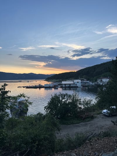 The image depicts a tranquil sunset scene over a large body of water in Brosh, Bulgaria. The sky transitions from a light blue with sparse cirrus clouds at the zenith to warm orange and yellow hues at the horizon, reflecting the setting sun which is partially visible as a bright glow behind a distant, dark-forested mountain range on the left. Scattered dark grey cumulus clouds are present in the mid-sky, particularly on the right.
The expansive body of water, likely a lake or reservoir, occupies the middle ground and reflects the sunset colors, creating shimmering patterns on its calm surface. Numerous small, modular floating docks and platforms, some with small structures or canopies, are distributed across the water. Several boats of varying sizes are moored to these structures; one white boat is visible through dense foliage in the lower-left corner, and another larger, white boat is situated among docks in the mid-ground.
Along the right bank, dense, dark green coniferous and deciduous trees cover a steep slope, extending from the water's edge upwards. Interspersed among the trees and along the shoreline are several white or light-colored buildings with dark roofs, appearing to be boathouses or lakeside establishments.
The immediate foreground on the left is characterized by lush green foliage, including trees and bushes, partially obscuring the water. On the lower right, an unpaved dirt path leads down towards the water. A silver sedan is parked on this path, partially concealed by foliage, and an empty, rusted metal frame, resembling a trailer chassis, rests on the ground nearby. The extreme lower right foreground consists of loose rocks, soil, and fallen branches, indicating an undeveloped or disturbed area. No people are visible in the scene.