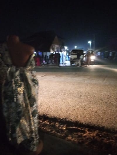A nighttime scene featuring an asphalt road stretching into the distance, illuminated by streetlights. A group of people stand near a building with a distinctive peaked roof, while a small truck and a motorbike are parked on the roadside. A person wearing a patterned garment stands in the foreground, partially obstructing the view. The overall atmosphere is dark, with the light sources creating stark contrasts and shadows.