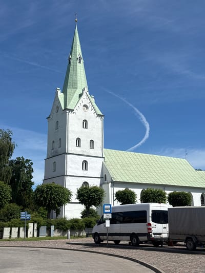 The image displays a white church with a green tiled roof and a tall, pointed steeple. The church has several arched windows and a clock face on its side. In front of the church, there is a white van and a trailer parked on a cobblestone street. Trees and bushes are visible on the side of the church and along the street. The sky is blue with some wispy white clouds, and a contrail forms an arc in the sky. The scene suggests it is a sunny day. Signs are visible on the side of the road, including a blue and white bus stop sign with a picture of a bus and the text "Tirgus laukums" (Market Square). Another sign appears to be a directional signpost. The overall impression is a view of a historical building in a European town. The image is oriented vertically.