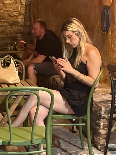 A blonde woman in a black strapless dress is seated on a green chair at a table outdoors, looking down at her mobile phone. She has blonde hair with lighter streaks, is wearing bracelets on her left arm, and has red nail polish. Next to her, to the left, a tan handbag rests on the table. Behind her, a man in a black t-shirt and grey shorts is seated, holding a glass of beer and looking to his right. The setting appears to be a courtyard with stone walls, consistent with the Old Town of Budva, Montenegro. A blue circular sign with white lettering is visible on the wall behind the woman.