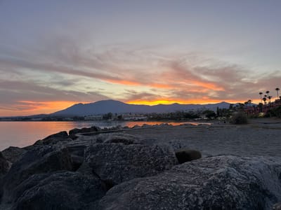 A serene coastal sunset scene unfolds in El Velerín, Spain. The sky is a canvas of soft purples, pinks, and vibrant oranges, with wispy clouds stretching across the horizon. In the distance, a range of mountains forms a dark silhouette against the glowing sky, with a town nestled along the coastline. The water is calm and reflects the warm hues of the sunset. In the foreground, large, dark rocks line the shore, interspersed with a sandy beach. A few palm trees are visible along the coastline, adding to the tropical ambiance. The overall impression is one of peaceful tranquility.