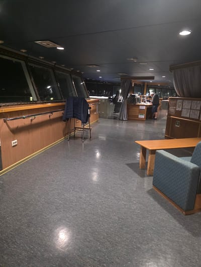 The image depicts the interior of a ship's bridge, likely at night, given the darkness visible through the large windows. The bridge is functional and appears to be in use. The floor is a dark gray linoleum. A dark blue jacket is draped over a metal stool positioned against a wooden paneled wall. A metal handrail runs along the wall. To the right, a wooden bench with blue upholstered seating is visible. In the background, a person in casual clothing is standing at a console or desk, appearing to be working. The ceiling is dark blue with several recessed lights. The overall impression is one of a working environment on a vessel. The image does not provide any specific visual cues to pinpoint its location to Brindisi, Italy.