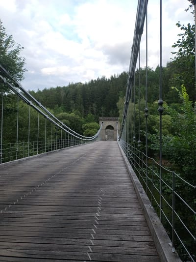 The image shows a wooden suspension bridge in Slavonice, Czechia. The bridge is made of weathered wooden planks and is supported by thick metal cables. The perspective is from the bridge, looking forward towards a stone archway at the far end. Dense green forest lines the sides of the bridge and extends into the background, under a cloudy sky. A body of water is visible to the right, partially obscured by trees. There are no people visible in the image. The scene appears to be an outdoor, natural environment with a man-made structure.