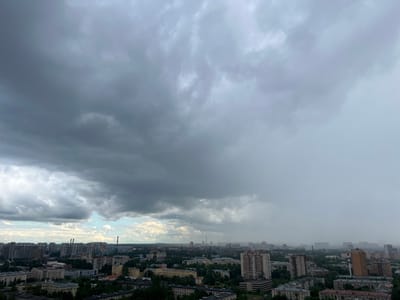 An aerial view of the city of Kirpichnyi Zavod, Russia, under a heavy, overcast sky. Dense, dark grey clouds dominate the upper portion of the image, with lighter, wispy clouds and hints of blue sky visible in the distance on the left. A visible downpour or heavy mist is occurring in the far distance on the right, obscuring the cityscape. The midground and foreground reveal a dense urban environment with numerous multi-story buildings of varying architectural styles, including residential apartment blocks and some industrial or commercial structures. Green trees are interspersed throughout the city, particularly in the lower foreground and between buildings. Chimneys of industrial facilities are visible on the horizon. The overall impression is of a cityscape preparing for or experiencing inclement weather. No people are visible in the image.