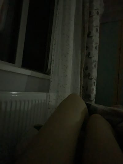 This image shows a close-up view of a person's legs. The person appears to be lying down, with their legs bent and partially obscuring a window or other flat surface. The lighting is dim, suggesting it is either nighttime or the room is very dark. The background consists of a textured surface with a repeating pattern of small, dark speckles on a lighter background. There is also a glimpse of what looks like a radiator or ventilation grate. The overall scene is dark and intimate, with the focus primarily on the form and texture of the person's legs. No specific activity or event is clearly depicted beyond the person resting. The location is identified as Zamostia, Ukraine.