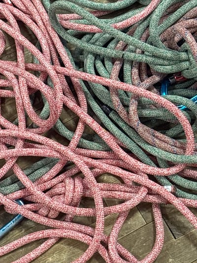 A cluttered arrangement of braided ropes, predominantly red-pink with white flecks and teal-green with multi-colored flecks, rests intermingled on a light brown wooden plank floor. Several metal carabiners, including blue and red-silver units, are partially visible among the tangled coils. The scene is static, depicting climbing or rigging equipment in disarray. This scene is located in the city of Tayshet, Russia.