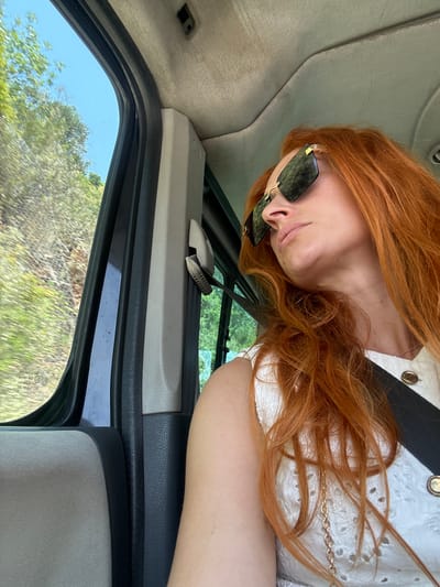 A young woman with long, wavy, ginger hair is resting her head on her arm inside a vehicle. She is wearing large, square, dark-tinted sunglasses with thin gold frames. Her left hand is resting on her cheek, and her right arm is visible in the lower right portion of the frame. She is wearing a white top with eyelet detailing and gold buttons. The interior of the vehicle appears to be grey and black. Through the open window, lush green trees and vegetation are visible against a bright blue sky, suggesting it is daytime and the weather is clear. The scene suggests a leisurely drive through a natural landscape. The image is rotated 90 degrees counterclockwise. No text is clearly visible.
