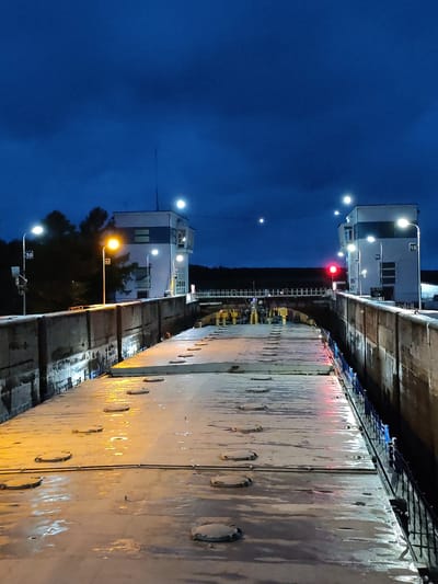 The image captures an outdoor scene at a ship lock in Devyatiny, Russia, during twilight. The sky is a deep blue with some cloud cover. On the left and right sides of the frame are imposing concrete structures forming the walls of the lock. The lock chamber itself is filled with a flat, metallic surface, likely the deck of a barge or a similar vessel, with several circular hatches visible. This surface is wet and reflects the orange glow of artificial lights.

Two prominent buildings, likely control towers for the lock, are situated on either side in the background. These structures are white with dark accents and are illuminated by multiple overhead lights. One tower on the right displays the number "10" and "11", and another on the left displays "10". A red traffic light is visible on the right-hand side, indicating a stop or caution.

Numerous streetlights, emitting a warm yellow light, are positioned along the lock walls and near the control towers, illuminating the scene. In the distance, silhouetted trees and the dark line of a forest can be seen against the twilight sky. The overall atmosphere is one of industrial functionality and the transition from day to night. There are no visible people or discernible actions taking place in the image.