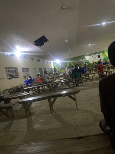 This image depicts a large indoor gathering space in Kubi A, Nigeria, illuminated by several overhead lights, one of which emits a strong glare. The ceiling, constructed with suspended acoustical panels, features a significant rectangular opening exposing a dark cavity above. Multiple ceiling fans are visible. The space is furnished with numerous long, wooden tables, each with integrated benches, arranged in rows across a concrete or hard-packed earth floor. Scattered plastic chairs in blue, green, and orange are interspersed among the wooden furniture.
Approximately two dozen individuals are visible, mostly seated at the tables, casually dressed. On the far wall, light-colored paint is partially obscured by decorative drapes in green and yellow fabrics. A large green banner with white text and diagrams, including outlines of human figures, hangs prominently on this back wall, suggesting a presentation or informational event. The left wall features barred windows and a wall-mounted fan. In the immediate foreground, the back and shoulder of a person wearing dark clothing are visible, indicating the photograph was taken from a seated perspective within the event.