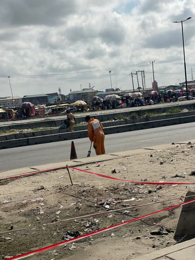 The image presents a busy roadside scene in Aiyetoto-Asogun, Nigeria, captured on what appears to be an overcast but bright day. The perspective is slightly elevated, looking across a multi-lane road towards a bustling market or commercial area.

In the immediate foreground on the right, there is a rough, unpaved patch of ground heavily littered with debris, including papers and plastic waste. Several red and white warning tapes or ribbons are strung across this area, possibly marking a boundary or indicating some form of work in progress. A small orange traffic cone is also visible.

A wide, dark grey paved road with multiple lanes stretches through the midground, separated by a concrete median strip that shows some overgrown vegetation. On the right side of this road, a person dressed in a bright orange, possibly reflective, uniform is visible from the back, appearing to push a cart or attend to a task. Another individual in a patterned dress stands nearby.

The left side of the road is densely populated and vibrant, suggesting a bustling market. Numerous people are gathered under a variety of colorful umbrellas, indicating stalls or temporary shelters for vendors. Vehicles, including cars and potentially motorcycles or auto-rickshaws, are visible amongst the crowd. In the background, various buildings, some multi-story, line the horizon, with several telecommunication antennae and utility poles rising above them. Tall street lights and an intricate network of power lines crisscross the sky, which is filled with large, diffused grey clouds.

The scene depicts active commerce and daily urban life. The bright, diffused lighting suggests it is daytime, likely midday or early afternoon. No specific text is clearly legible on any signs or banners in the image.