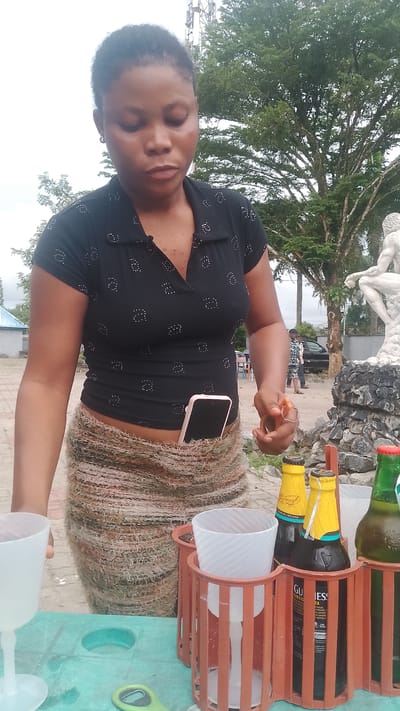 A woman stands outdoors, her head bowed and eyes closed as if in thought or prayer. She is wearing a black polo shirt with a subtle white dot pattern and a patterned, textured skirt. A smartphone is tucked into the waistband of her skirt. In front of her, on a green table, are two white plastic cups, one in a wooden holder with an orange tint, and another in a similar holder with two bottles of Guinness Foreign Extra Stout and a green bottle of Guinness. To the right, another green bottle is partially visible. Behind her, a large tree with lush green foliage dominates the background, with a white statue of a person on a rocky base to the right. In the distance, a utility pole and a building can be seen, along with other people and a car. The sky is overcast, suggesting a cloudy day. The scene appears to be set in Agbarho, Nigeria. The woman's expression and posture suggest a moment of quiet contemplation amidst an outdoor setting, possibly a roadside vendor or a gathering.