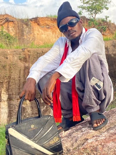 This outdoor portrait features a man posing against a rugged, rocky backdrop, likely in Afikpo, Nigeria. He has dark skin and is styled in a black beanie, dark sunglasses, a white long-sleeved button-up shirt with rolled cuffs, and loose-fitting gray pants. A bright red scarf or sash is draped around his neck, providing a striking color contrast. He completes his look with black slides.
The man is positioned with a confident lean, looking directly towards the camera. His right hand holds the handle of a stylish black handbag. The bag is textured with geometric patterns and features gold-colored accents, including the prominent text "BRANDON JOE" and a circular "BJ" logo.
The setting appears to be a natural landscape with steep, earthy-toned rock formations and sparse green vegetation, suggesting a cliffside or quarry environment. The sky is visible in the upper left, indicating daylight conditions and sunny weather. A small white tag is visible on the gray pants, displaying the word "ROHV" and some other indistinct text. The overall scene suggests a casual fashion photoshoot against a dramatic natural setting.