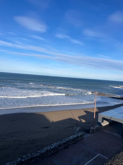 The image presents a wide view of the beach and adjacent ocean in Zumaia, Spain. The upper two-thirds of the frame are dominated by a clear blue sky with several wispy, elongated white clouds. Below the horizon, a deep blue sea exhibits multiple parallel wave formations, with white foamy surf breaking onto a dark sandy beach. A single, small white sailboat is discernible on the distant ocean surface.

The lower third of the image shows a portion of the sandy beach, which appears darker due to shadows cast from structures located to the left, outside the frame. Scattered footprints are visible on the shadowed sand. In the foreground, a low stone wall, constructed from grey and brown stones, runs horizontally, separating the beach from a reddish-brown paved area. A set of stone steps descends from this paved area towards the sand. To the right, a modern building with a flat, light-colored roof and dark walls is partially visible at the edge of the beach. A tall, slender, reddish-brown pole stands vertically on the sand directly in front of this building. Exposed rocky formations are visible along the waterline on the far right side of the image. No individuals or animals are present in the scene.