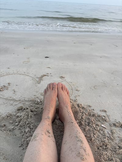 A close-up, low-angle shot shows a person's legs and feet partially buried in sand on a beach. The sand is light brown and wet near the water's edge. A heart shape has been drawn in the sand in front of the person's feet. The ocean stretches out in the background with gentle waves breaking on the shore under a pale sky. The scene appears to be a tranquil setting by the sea.