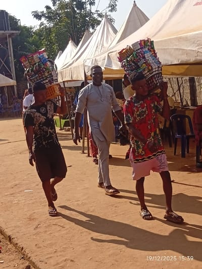 The scene depicts an outdoor gathering, reportedly in Isinyi-Nando, Nigeria, captured on 19/12/2025 at 15:39. Three males walk from left to right across an unpaved, reddish-brown dirt surface under direct sunlight, casting distinct shadows.
The foreground features two young males carrying tall stacks of packaged snack items. The male on the left wears a black t-shirt with a white graphic, black shorts, and white flip-flops; he carries a light-colored plastic crate filled with snacks on his left shoulder. The male on the right wears a multi-colored patterned short-sleeved shirt, light pink shorts, and dark slides; he carries a dark woven basket filled with snacks on his right shoulder.
Between these two, slightly behind, an adult male walks, wearing a light grey tunic, light grey trousers, and brown sandals. He has dark-framed glasses and a silver-colored cross pendant on a chain around his neck.
In the background, multiple white, peaked tents are erected, indicating an event or temporary market setup. Blue plastic chairs are visible under or near the tents, with some indistinct figures present. On the far left, a large black speaker tower is partially visible. Green foliage from trees is seen above the tents against a bright sky.