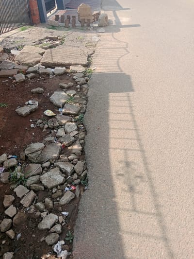 A sidewalk is severely damaged with broken concrete pieces scattered on the dirt ground. The adjacent asphalt road appears in good condition, with shadows of a fence cast across its surface. In the background, there are several stone carvings. Debris litters the area between the sidewalk and the road. The scene suggests neglect and disrepair of the pedestrian infrastructure.