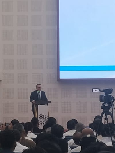 A man in a suit and tie stands at a podium, speaking to an audience. The room has a tiled wall and a large screen displaying a light blue background with a horizontal blue line. A camera on a tripod is positioned in front of the audience. The audience members are mostly seated, some are visible in the foreground, and others are further back. The overall scene appears to be a presentation or conference.