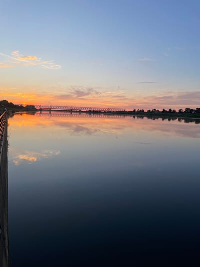 This is a vertical, eye-level, medium shot of the Sovetsk Railway Bridge over the Neman River at sunset. The sky is a gradient of soft blues, pinks, and oranges, with wispy clouds reflecting on the calm water. The bridge, a prominent steel structure with intricate lattice work, stretches across the horizon. The far bank of the river is lined with trees and some distant buildings, all silhouetted against the colorful sky and mirrored in the water. On the left side of the frame, a dark, textured wall or embankment with a railing runs along the edge of the river. A single person is visible on the embankment, looking towards the sunset. The overall mood is serene and picturesque, capturing the beauty of the natural landscape and the industrial element of the bridge at dusk.