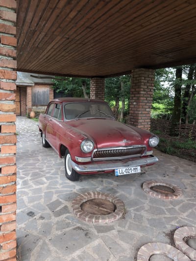 In Siktarva, Georgia, a vintage red Volga GAZ-21 car is parked under a rustic wooden and brick shelter. The car, covered in a light layer of dust, features a chrome grille and white-rimmed wheels. The ground is paved with irregular flagstones, and three circular brick structures with wooden lids are visible on the ground near the car. In the background, a wooden building with a sloping roof stands, and beyond that, lush green trees and a woven fence are present. The scene depicts a tranquil, outdoor setting with a focus on the classic automobile.
