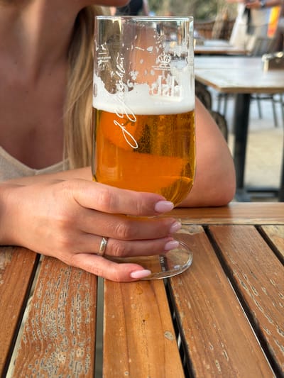 In a medium shot, a person's hands and a glass of beer are prominently featured on a weathered wooden table. The person, who appears to be female with blonde hair, is wearing a light-colored top and a silver ring with small diamonds on her left ring finger. Her nails are painted a light pink. She is holding a tall glass filled with amber-colored beer, topped with a frothy white head. The glass has some writing on it, including what appears to be a crest and cursive text. The background is blurred, showing other tables and people in what looks like an outdoor seating area, possibly a cafe or restaurant. The lighting suggests it is daytime, with the sun casting a warm glow. The overall scene evokes a casual, relaxed atmosphere. The location is identified as the city of Zagora, Montenegro.