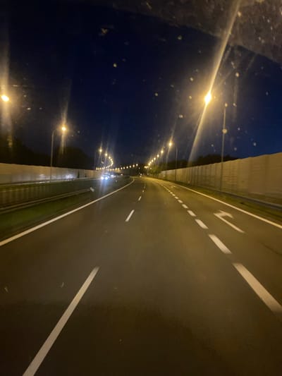 This image depicts a nighttime view from a vehicle traveling on a highway in the vicinity of Kokotów, Poland. The road is illuminated by overhead streetlights, creating a clear path ahead. The highway has multiple lanes, separated by white lines. On the right side of the road, a sound barrier made of beige panels is visible. In the distance, other vehicles with illuminated headlights can be seen. The sky is dark, indicating it is nighttime. The image is taken from inside a vehicle, likely through the windshield.