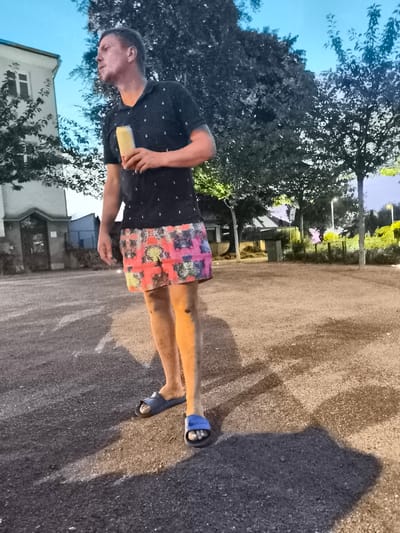 A man stands outdoors in the city of Kolín, Czechia, holding a can and wearing a dark polo shirt with a pattern of small anchors and colorful, patterned shorts. He is also wearing blue slides. The man is standing on an asphalt surface next to a gravel area. In the background, there is a building, trees, and streetlights. The sky is a twilight blue.