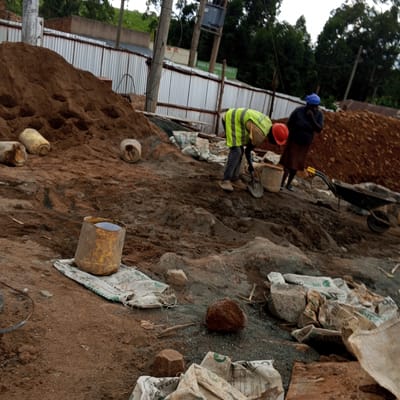 The image shows a construction site in Keyian Ward, Kenya. Two people are present, one wearing a bright yellow vest and a red helmet, digging into a mound of dirt with a shovel. A second person, wearing a blue head covering and a dark jacket, stands nearby. A wheelbarrow rests on the right side of the scene, partially filled with material. The area is covered with piles of dirt and construction materials, including several sacks, rocks, and buckets. A corrugated metal fence is visible in the background. Utility poles with electrical equipment are also visible behind the fence.