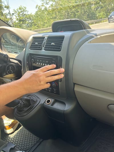 The image shows the interior of a car, likely a Fiat, in Golubovići, Montenegro. A person's hand, with manicured fingernails, is resting on the car's radio and climate control panel. The hand is positioned over several buttons and knobs. The dashboard features air vents, a car stereo with several buttons and a display screen, and controls for the car's climate. The "AIRBAG" text is visible on the dashboard. The scene appears to be set during the daytime, with sunlight filtering through the trees visible outside the car's windows, suggesting a warm and possibly sunny day. The overall mood is casual, with the focus on the car's interior and the hand interacting with the controls. The presence of a gear shift lever and a key in the ignition indicates the car is parked or in neutral. The image is taken from the perspective of someone sitting in the driver's seat.