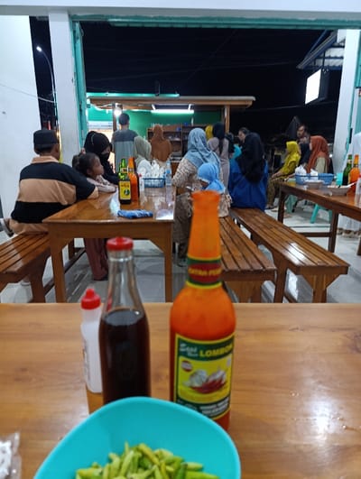 The scene is a night-time view from a table at a restaurant or food stall. There are several people sitting at tables in the background. A group of people are seated at a wooden table in the foreground with two bottles of sauces, a bowl of green beans, and a blue bowl. Two bottles of chili sauce, one red and one brown, are visible in the foreground. The table is made of wood with a shiny surface. The restaurant appears to be located in Kecamatan Asembagus, Indonesia, at the coordinates -7.75 latitude and 114.21 longitude. The environment is outdoor with a dark sky and a few lights. The scene is casual and typical of an Indonesian restaurant.