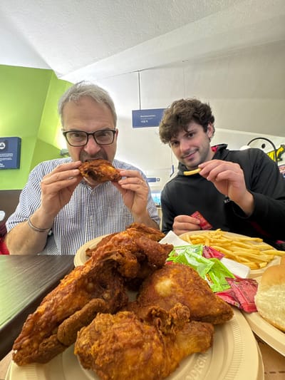 Two men pose with a feast of fried chicken and french fries. The older man with glasses holds a chicken wing, while the younger man eats a fry. A plate overflows with golden-brown fried chicken pieces. Next to it, a separate plate holds a mound of fries, packets of condiments, and buns. The photo is taken at an angle, with white walls and blue signs in the background.