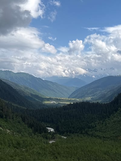 The image displays a panoramic view of a mountainous valley in Mazeri, Georgia, under a partially cloudy sky. The foreground is dominated by dense evergreen and deciduous forests covering the slopes of steep, verdant mountains. A winding river or stream is visible in the lower middle section of the image, cutting through the trees. The mid-ground reveals a wider valley floor with agricultural fields or meadows, and a settlement or village with scattered buildings. In the background, a range of majestic, snow-capped mountains rise, partially obscured by clouds. The overall atmosphere is serene and natural, showcasing the rugged beauty of the Georgian landscape. There are no people or animals visible in the image, and no specific actions or interactions are depicted. The scene captures a natural environment with elements of human habitation in the distance.