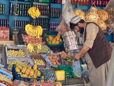 A man wearing a brown vest and long pants is standing at a market stall in Dand, Afghanistan (31.618, 65.628). The stall is filled with fresh produce including bananas, potatoes, and papayas. The man is holding a bag of okra, which is on display in the foreground. The stall is located on the right side of the image. To the left of the stall, the shelves are stocked with a variety of items, including canned goods, drinks, and bags of dried fruits. The shelves are also decorated with hanging bananas. The walls are painted blue, and there is a sign above the shelves that reads "Dand, Afghanistan."