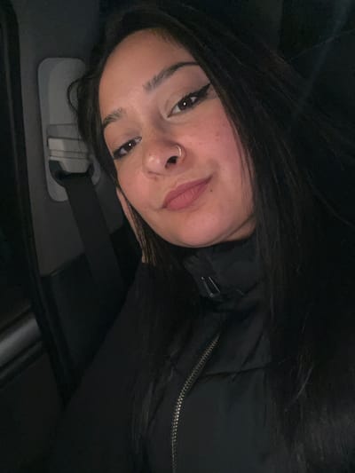 A young adult female is depicted in a close-up portrait, positioned inside what appears to be a vehicle. She has long, dark hair, brown eyes, and a light-to-medium complexion with visible freckles across her nose and cheeks. Eyeliner is applied to her upper eyelids, and her lips are colored with a light pink lipstick. A silver nose ring is present on her septum. She is wearing a black long-sleeved garment underneath a black puffer vest with a silver zipper running down the front. Her right hand is positioned near her jawline. She gazes directly at the camera with a subtle, closed-mouth smile. In the blurred background to the left, a grey car seatbelt buckle and strap are visible. The environment is dark and dimly lit, consistent with a vehicle interior at night. The specific location of Gerakas Municipal Unit, Greece, cannot be ascertained from the visual evidence within the image.