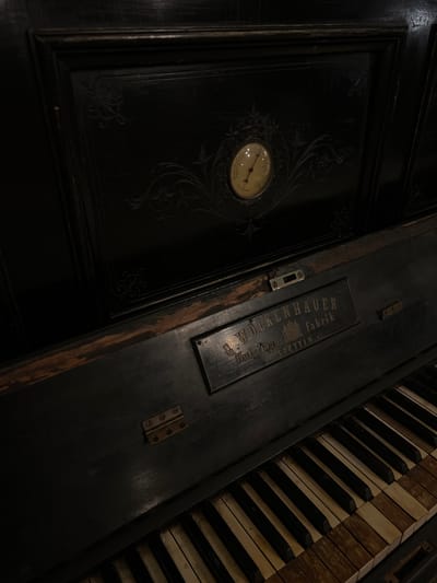 A close-up, dimly lit, high-angle shot focuses on the upper portion of an antique black piano. The piano features ornate carved details on its dark wood casing, with a small, round thermometer embedded in the center of the upper panel. A brass plaque with the inscription "C. WÖLKENHAUER Hof-Piano-Fabrik Stettin" is affixed to the front edge of the piano, above the keyboard. The keys of the piano are visible, showing a mix of black and yellowed ivory or bone, some appearing worn and discolored. Hinges are visible on the left side of the piano. The overall aesthetic suggests a vintage and possibly neglected instrument, evoking a sense of history and quiet elegance. The scene is devoid of any people or discernible action, presenting a static, object-focused composition. The location in Beit Hanan, Israel, is not evident from the visual elements within the image.