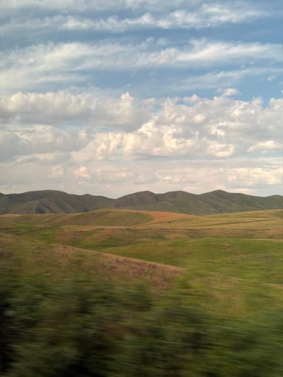 The image depicts a vast, rolling landscape under a cloudy sky. The foreground is blurred, suggesting motion, and shows patches of green and brown vegetation, possibly foliage from a vehicle's passage. The mid-ground consists of undulating hills covered in a mix of green and golden grasses. The background features a series of larger, darker green hills that fade into the distance. The sky is a pale blue with numerous horizontal streaks of white clouds, some wispy and others more dense. The overall scene conveys a sense of open space and natural beauty. The location is identified as Karagai Bulak, Kyrgyzstan.